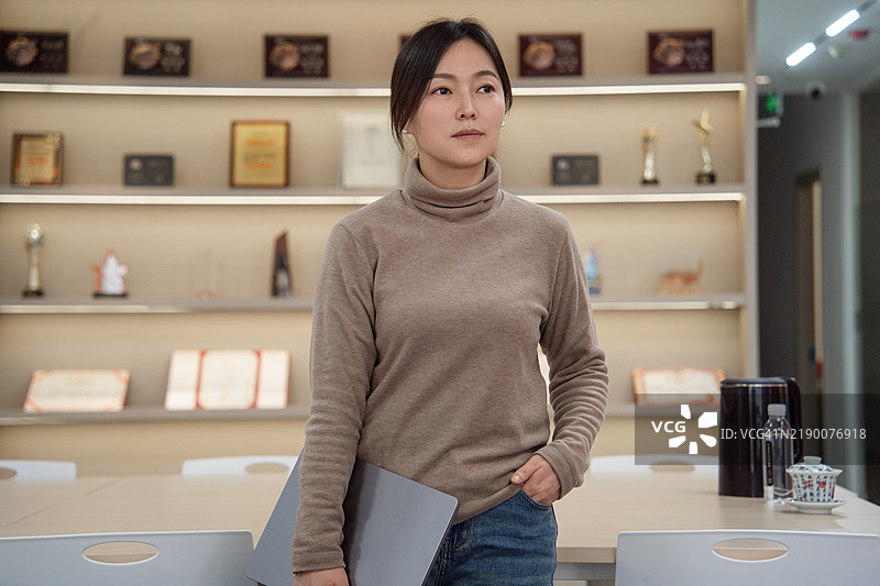 一位自信的亚洲女性，手持笔记本电脑，站在荣誉墙前，目光远眺。图片素材