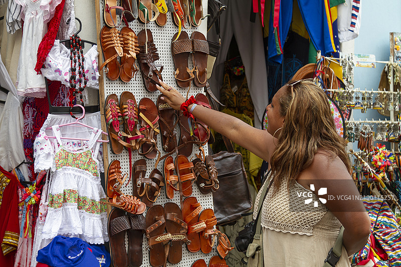 一位非洲裔成熟的女性背包客在巴西巴伊亚州萨尔瓦多的佩洛里尼奥色彩缤纷的纪念品商店里挑选凉鞋。图片素材