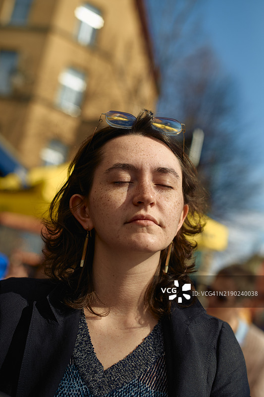 一位年轻女性闭着眼睛，头戴太阳镜，站在户外享受阳光。图片素材