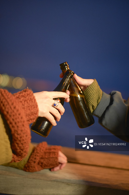 两个人在户外黄昏的环境中碰撞棕色啤酒瓶，背景为蓝色，比利时图片素材