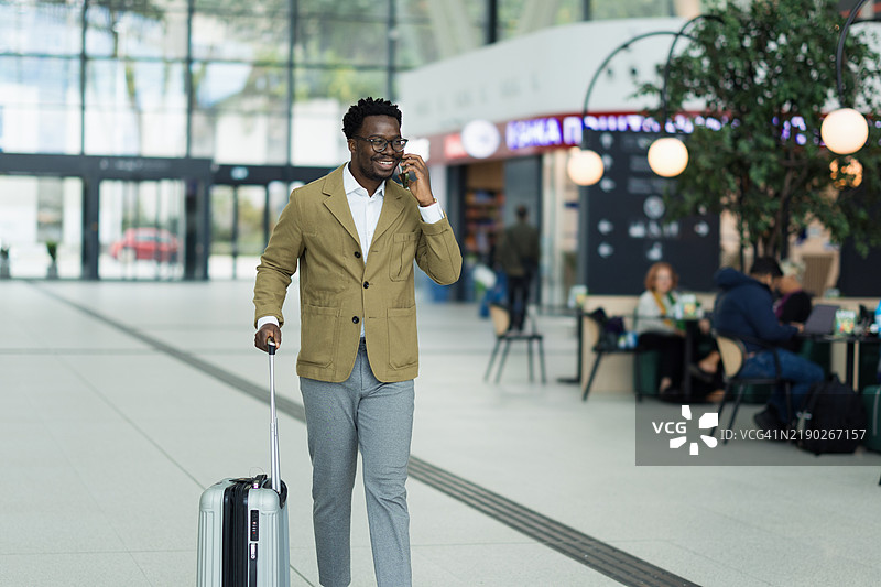 在机场航站楼内使用智能手机的商人，同时拉着行李。图片素材