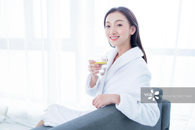 一位穿着白色浴袍的女性正在喝一杯加柠檬的水。图片素材