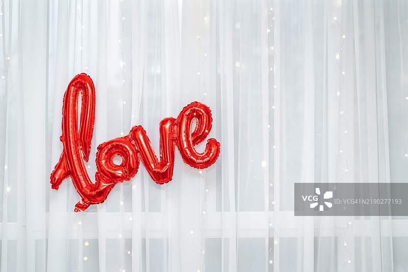 一个鲜艳的红色气球呈现出“爱”字的形状，悬挂在柔和的白色窗帘前，为室内的情人节庆祝活动营造了浪漫的氛围。图片素材