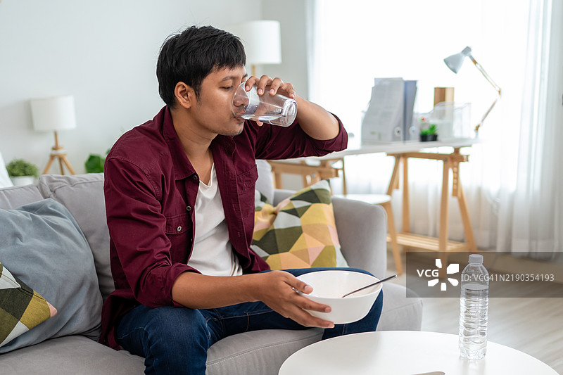 亚洲年轻男子在家喝水和吃方便面。迷人的男子坐在沙发上，感到快乐和放松，享用健康的早餐，享受客厅里的美食时光。图片素材