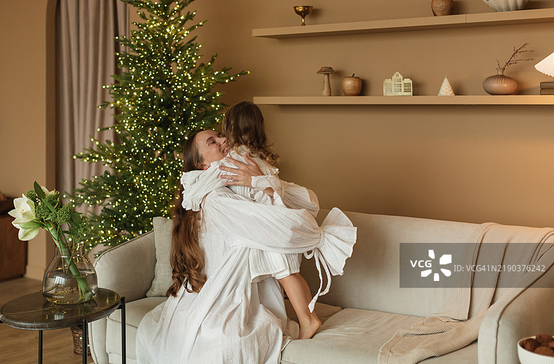 快乐的母女穿着睡衣坐在沙发上，紧紧相拥，旁边是一棵闪亮的圣诞树。图片素材