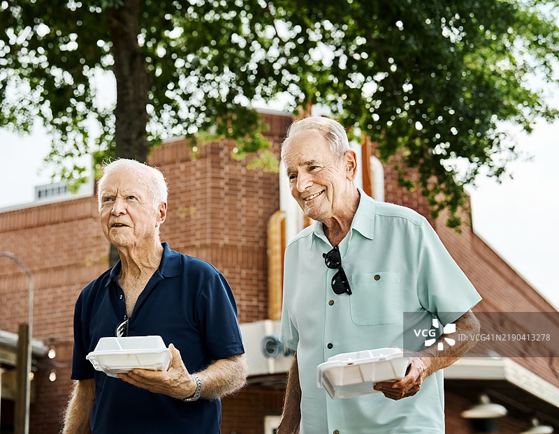 两位快乐的老年男性朋友在户外走着，手里拿着外卖食物图片素材