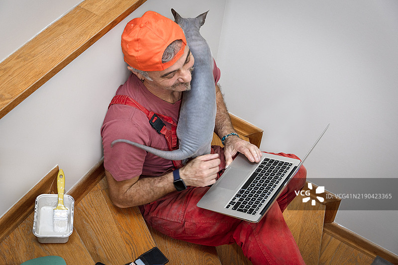 房屋油漆工在休息时用笔记本电脑，肩膀上有只猫。图片素材