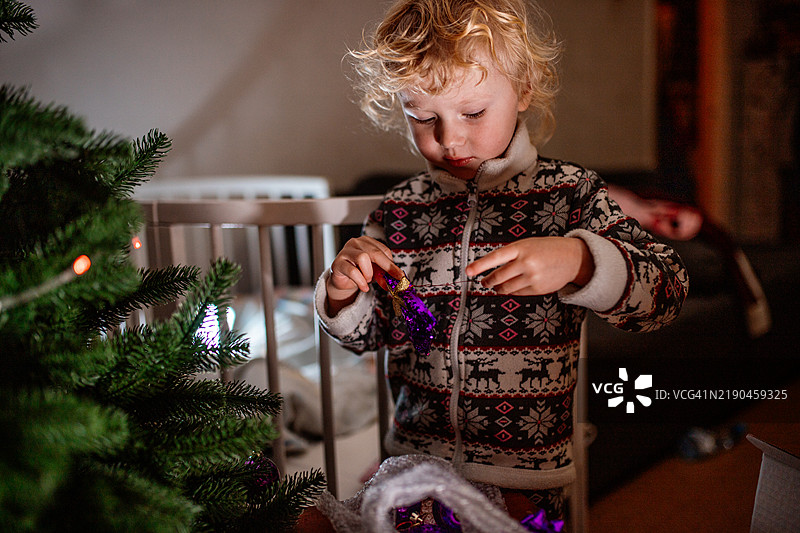 穿着圣诞睡衣的小女孩快乐地用紫色装饰品装饰圣诞树。图片素材