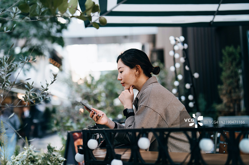 年轻的亚洲女性在街边咖啡馆放松，周围环绕着绿色植物，正在用智能手机发短信和浏览社交媒体。享受着一个悠闲的下午时光。生活方式、休闲与科技图片素材