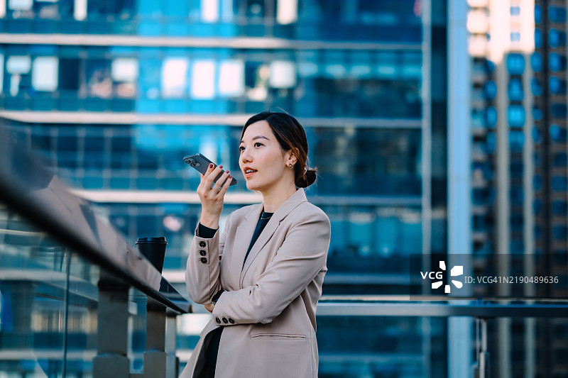 年轻的亚洲女商人在屋顶上与智能手机上的虚拟助手ChatGPT AI聊天应用进行对话，背景是现代摩天大楼。聊天机器人对话。互联网虚拟助手。人们、人工智能、创新与科技。图片素材