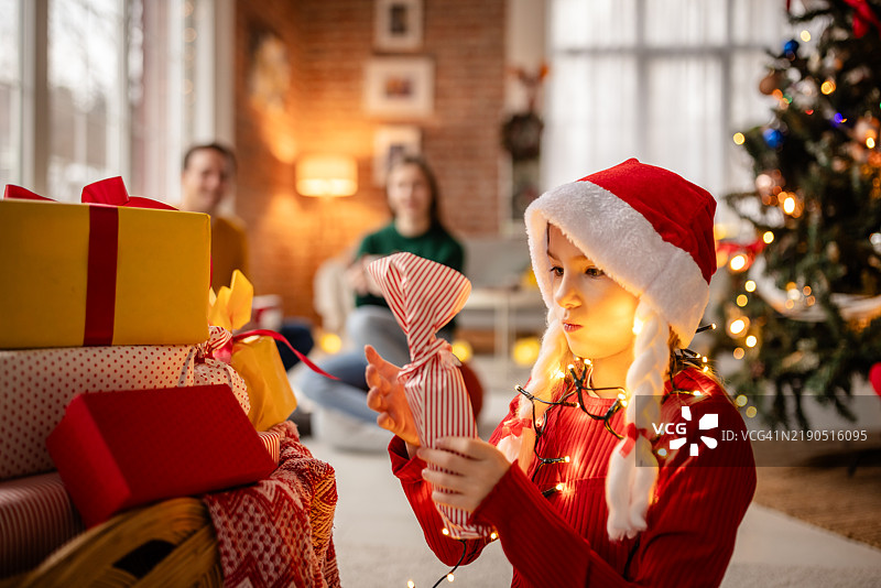 孩子在温馨的家中享受打开圣诞礼物的乐趣，周围有装饰好的圣诞树和家人。图片素材