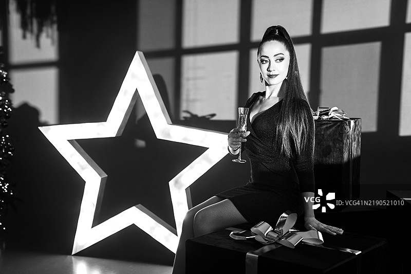 一位穿着优雅黑色连衣裙的女性坐在一个大礼物上，背景是星星，手中拿着一杯香槟。图片素材