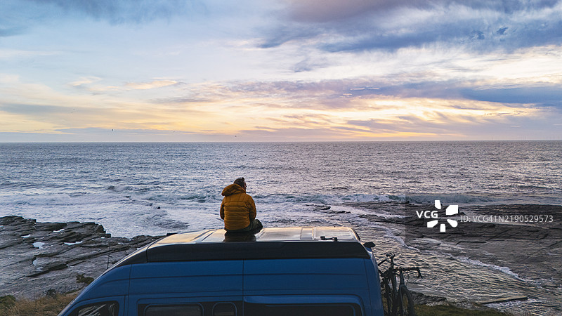 一位成熟的男子坐在他的房车顶上，等待海上的日出。图片素材
