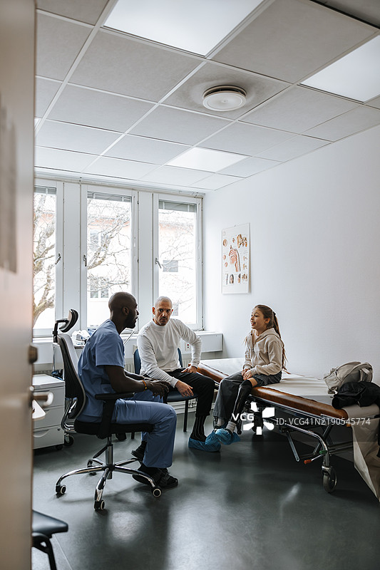 女孩与父亲坐在床上，正在与男性医生讨论，地点是检查室。图片素材