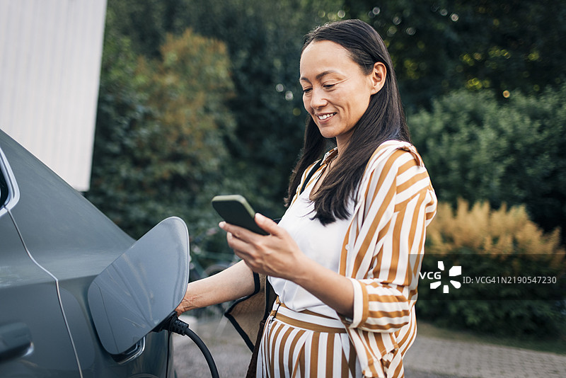 微笑的成熟的女性在后院给汽车充电时使用智能手机图片素材