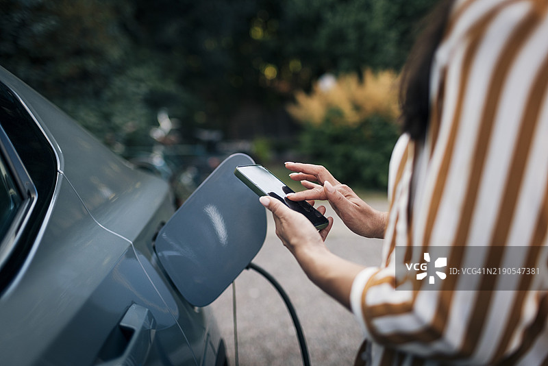 中年女性在后院为汽车充电时使用智能手机的中部特写图片素材