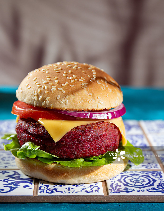 两款色彩鲜艳的甜菜汉堡，上面放有融化的奶酪、脆嫩的生菜、多汁的番茄和新鲜的洋葱，夹在芝麻种子面包中。蓝色背景。图片素材