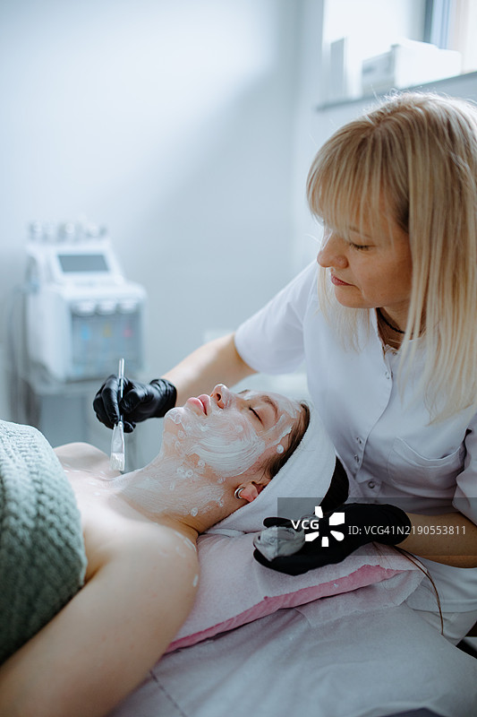 护肤程序。美容师的手在面部护理中小心地涂抹奶油面膜。水疗护理。图片素材