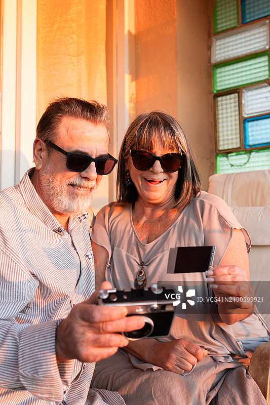 一对恩爱老年夫妇在日落时分的浪漫约会上，使用胶卷相机拍摄即时纸质自拍照。图片素材