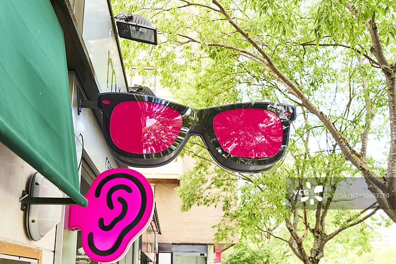 从低角度正面拍摄的眼镜店外墙上的标志，外面有红色镜片，背景中有一棵树。图片素材