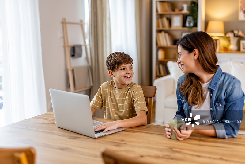 母亲和儿子在家餐厅一起用笔记本电脑工作，分享温暖的微笑图片素材