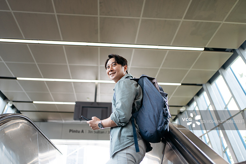 在城市地铁站内，手持智能手机的亚洲背包客正在乘坐自动扶梯。图片素材