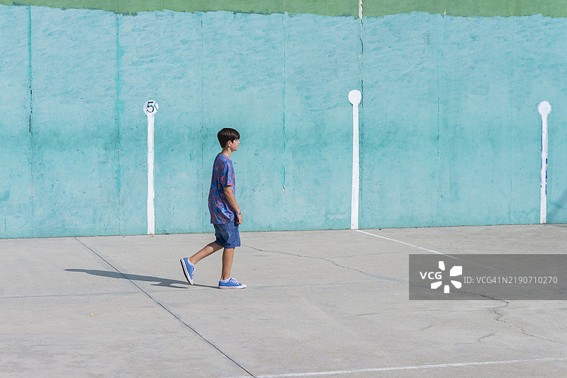 阳光明媚的日子里，穿着短裤和T恤的快乐男孩在前面的网球场上走动。图片素材