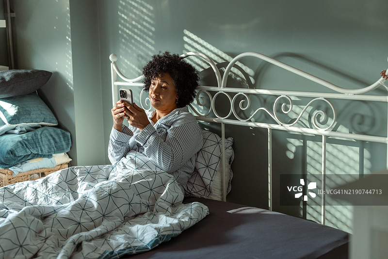 早晨的阳光下，女人坐在床上使用智能手机图片素材
