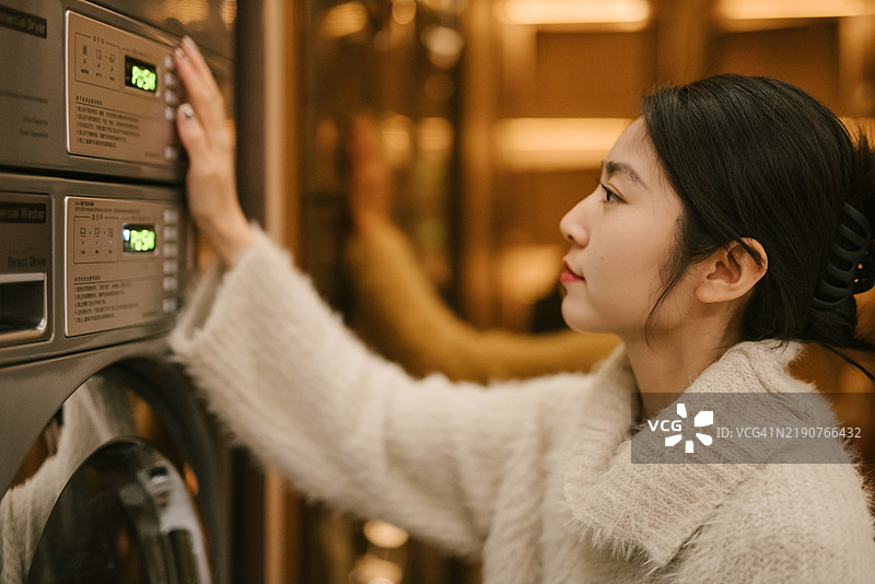 一位20多岁的年轻亚洲女性在光线明亮的洗衣房里操作洗衣机。图片素材