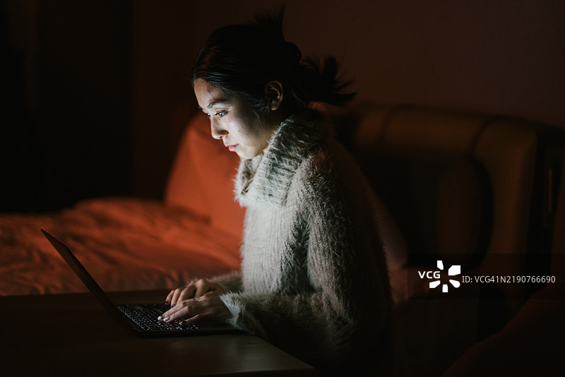 一位年轻的亚洲女性，可能二十多岁，专注于她的笔记本电脑，身处昏暗的房间，暗示着深夜工作或学习。图片素材
