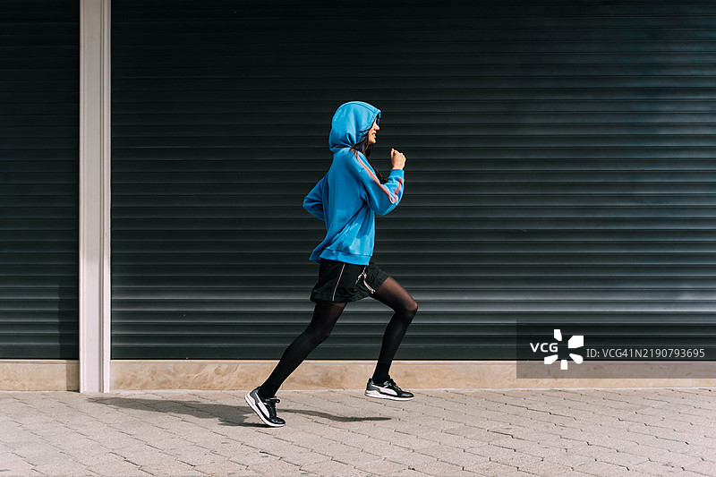 一位穿着蓝色连帽衫的年轻亚洲女性在街道上沿着黑色墙壁奔跑。生活方式与青年文化。图片素材