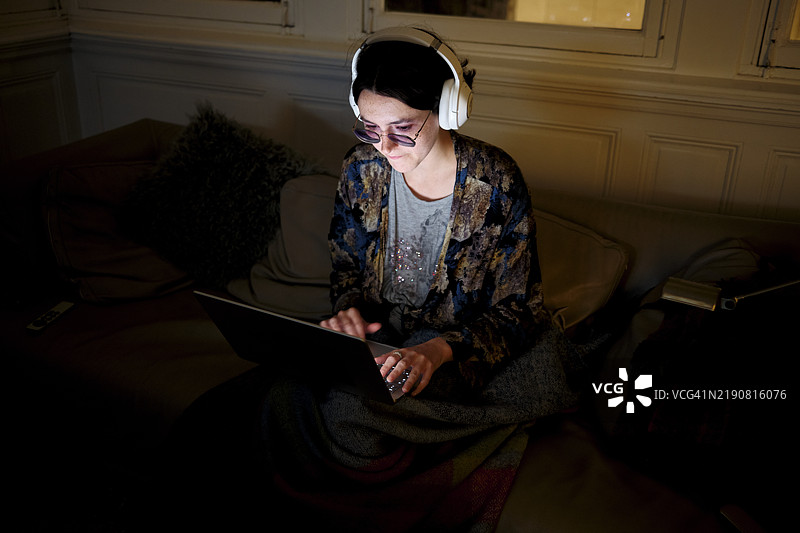 戴着耳机的女性专注于昏暗房间里的笔记本电脑屏幕图片素材
