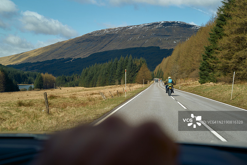 苏格兰高地，一条风景如画的公路，摩托车手在松树和山脉环绕的道路上行驶，蓝天晴朗。图片素材