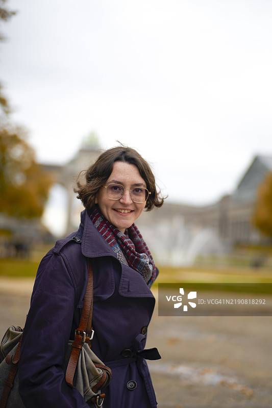 微笑的女性穿着紫色外套，围着围巾，站在历史遗迹前，天空多云。图片素材