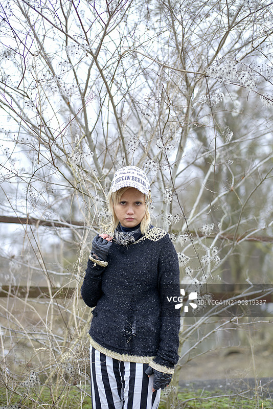 年轻女性穿着条纹裤和针织毛衣，戴着冬帽，站在光秃的树木之间，东德图片素材