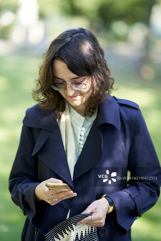 一位戴眼镜、穿着海军蓝外套的女性专注于她的智能手机，站在户外，背景是树木，德国德累斯顿。图片素材