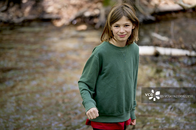 穿着绿色毛衣的男孩站在德国勃兰登堡的一条林间溪流旁，周围有落叶。图片素材