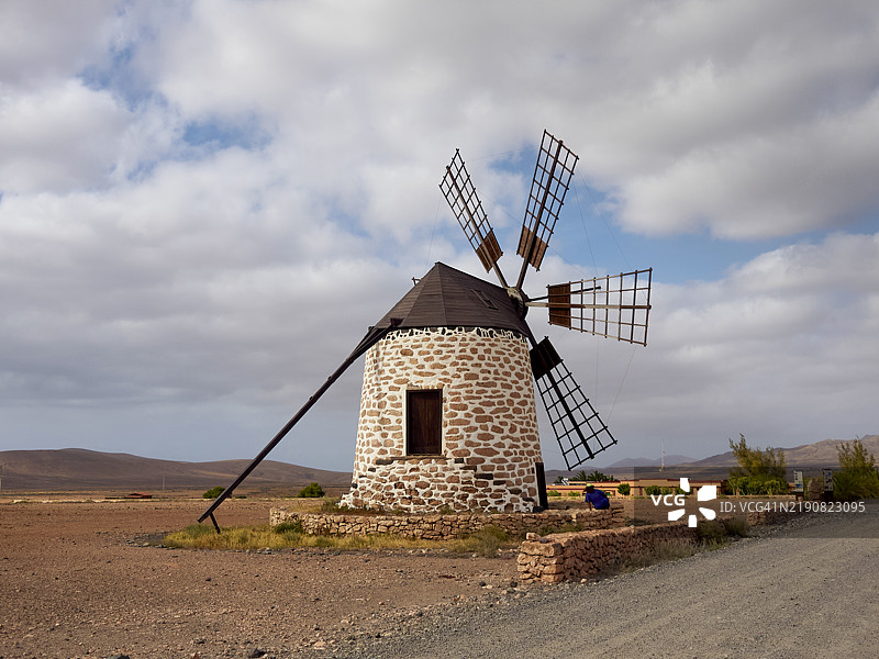 传统石制风车，木制风扇在多云的天空下，旁边是一条碎石路，背景是一个村庄，位于西班牙富埃特文图拉岛。图片素材