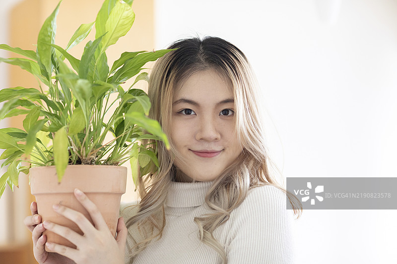 微笑的年轻女性在室内抱着一盆植物，背景明亮。图片素材