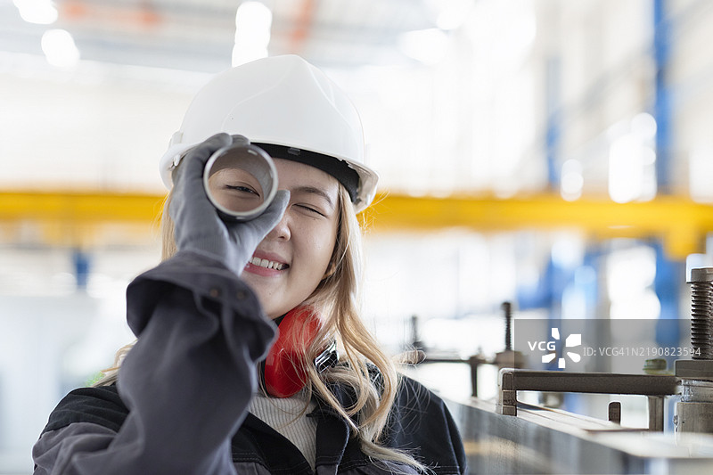 微笑的女性工程师戴着安全帽，在工业环境中透过金属杆观察，德国图片素材
