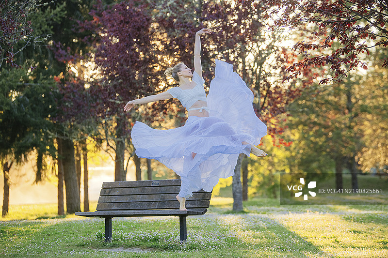 芭蕾舞者在日落时分于公园长椅上优雅地摆出一个姿势，背景是盛开的树木。图片素材
