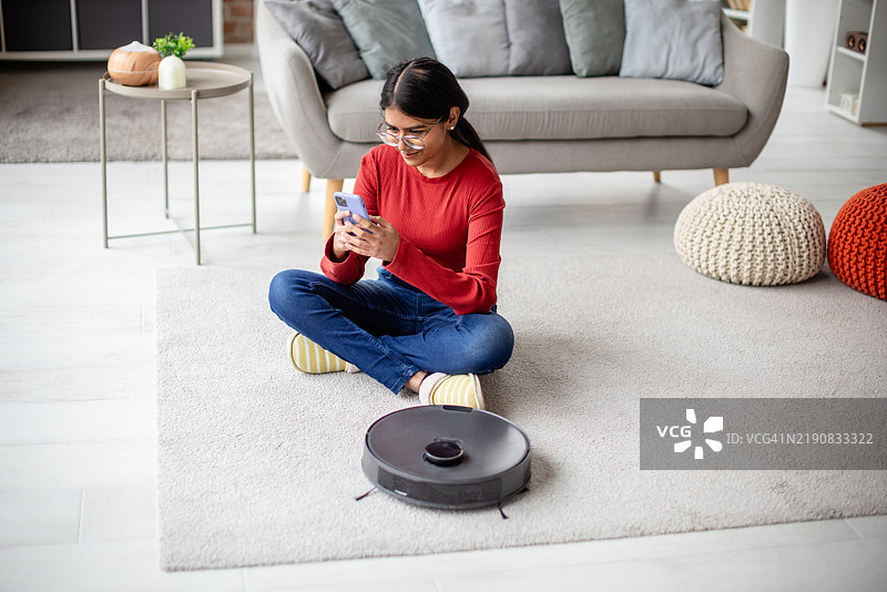 微笑的女性通过智能手机与智能吸尘器互动图片素材