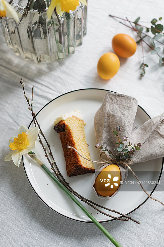 复活节餐桌布置的俯视图，包含复活节彩蛋、折叠成兔耳形状的餐巾、树枝、黄水仙花和一片涂有黄油霜的蛋糕。图片素材