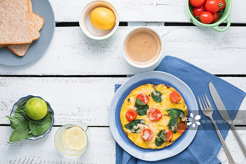 从上方俯视一份奶酪、樱桃番茄和菠菜煎蛋卷，旁边有一杯卡布奇诺和柠檬水，放在木桌上。图片素材