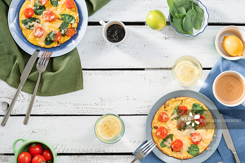 俯视两份奶酪、樱桃番茄和菠菜煎蛋卷，旁边有一杯卡布奇诺、一杯黑咖啡和一杯柠檬水，摆放在木桌上。图片素材