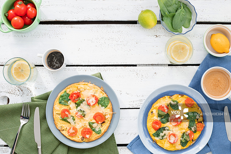 俯视两份奶酪、樱桃番茄和菠菜煎蛋卷，旁边有一杯卡布奇诺、一杯黑咖啡和一杯柠檬水，摆放在木桌上。图片素材