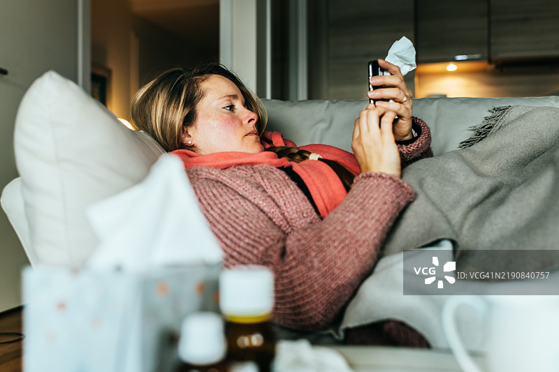 一位女性躺在沙发上，使用手机写病假条，感冒并感到不适。图片素材