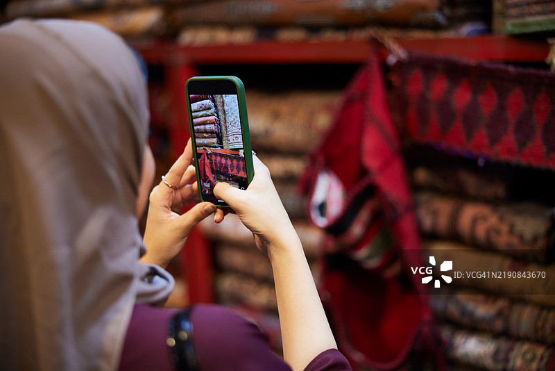 这位女士手持智能手机，在市场上选择最好的地毯时拍照。图片素材