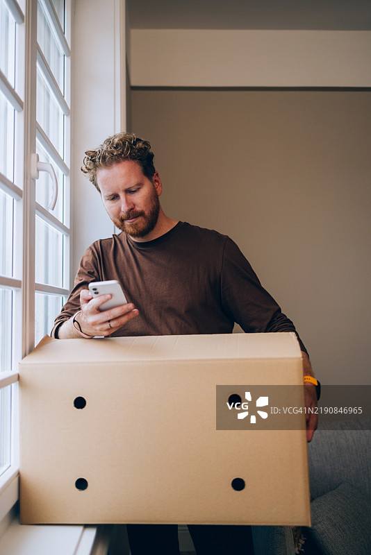 男人在明亮的窗户旁边用智能手机拿着一个纸箱图片素材