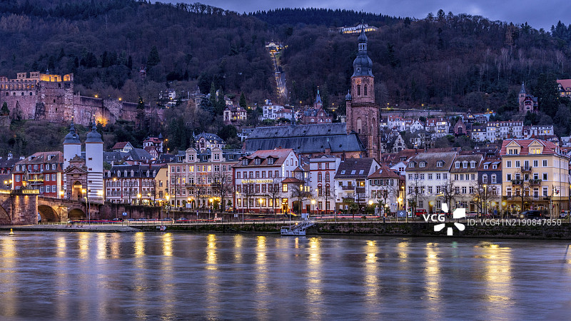 海德堡历史悠久的老城区，黄昏时分灯光照亮的建筑和前景中的内卡河，海德堡内卡河畔的步道图片素材